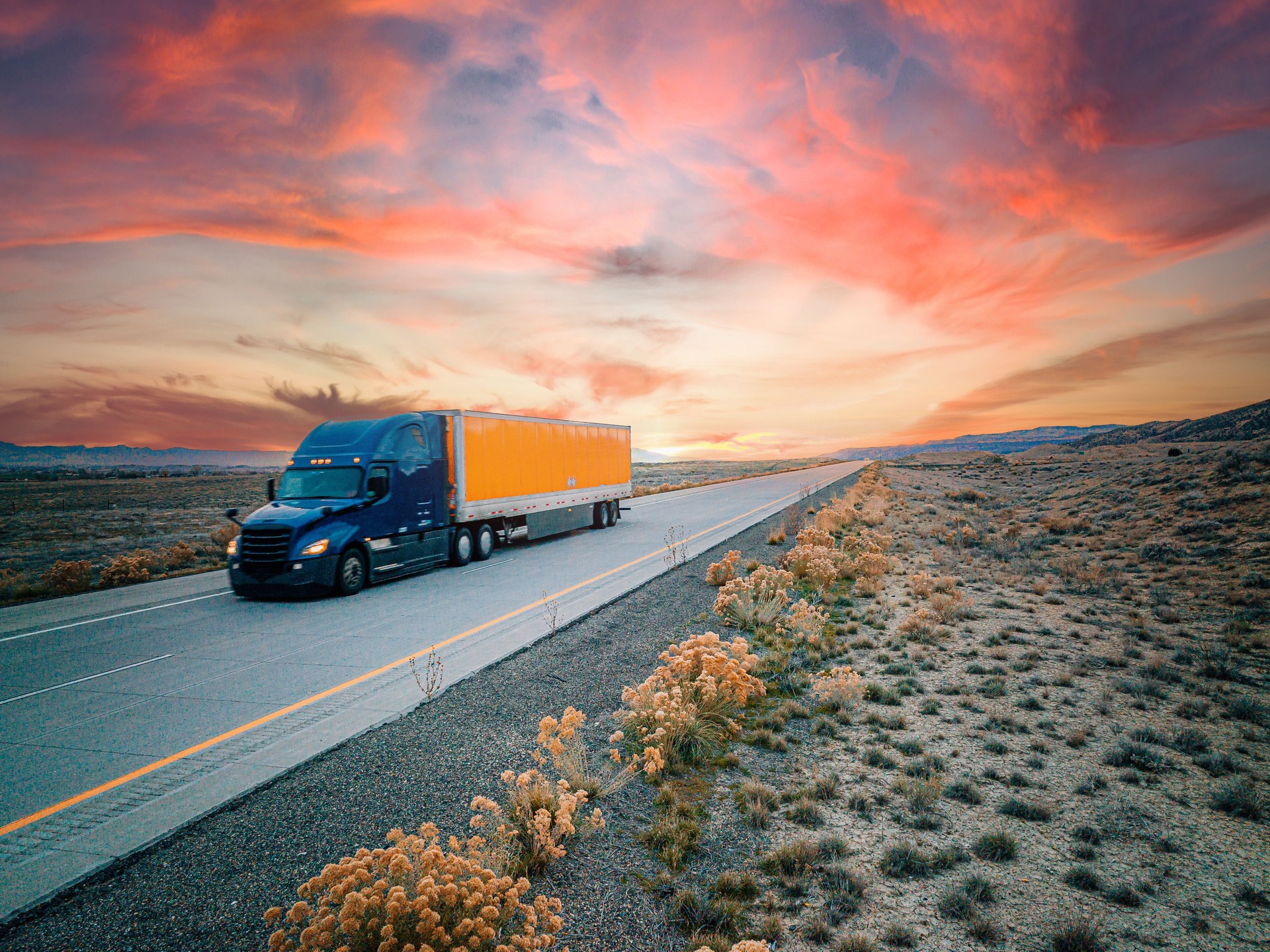 Low Angle Aerial Drone Shot of a Blue and Orange Semi Truck on the Highway with a Colorful Sunset